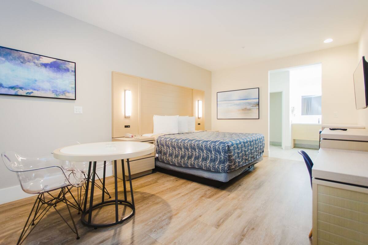 A modern hotel room with a bed, a round dining table, and a chair. There are two wall-mounted lights and a large window.