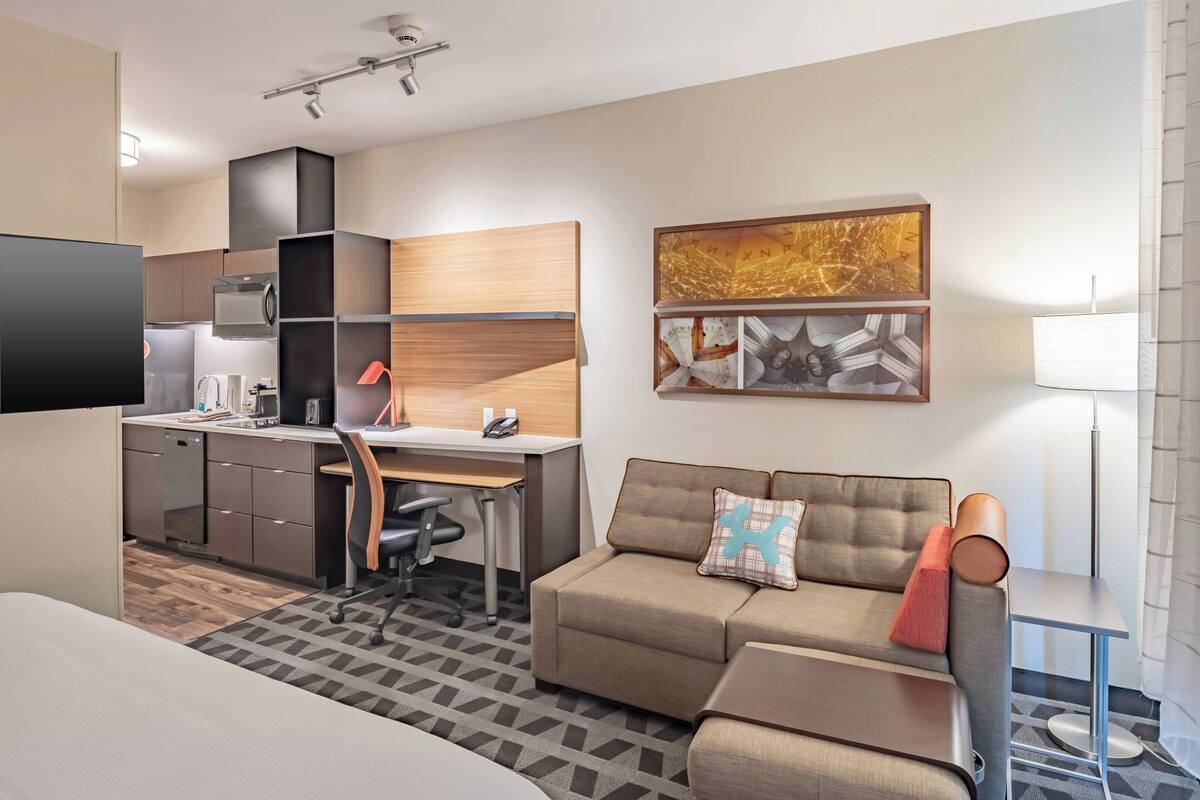 A modern hotel room with a kitchenette, a sofa, a desk, and a flat-screen TV.
