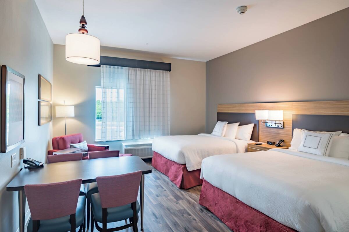 A hotel room with two beds, a desk with chairs, a table, and a window with curtains.
