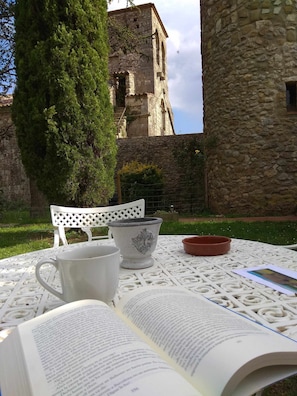 Un livre ouvert est posé sur une table blanche à l’extérieur, à côté d’une chaise blanche, d’un pichet blanc et d’un bol rouge. On aperçoit une tour de pierre au fond.