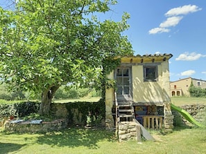 Un bâtiment rustique en pierre, doté d’un petit escalier en bois menant à une porte, le tout entouré de verdure et sous un ciel dégagé.