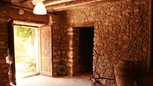 Um quarto rústico com paredes de pedra, uma porta de madeira, um grande vaso de barro e um lustre pendurado.