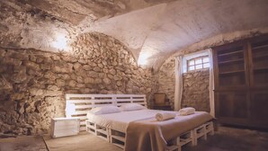 Quarto rústico com paredes de pedra, uma cama de madeira, uma mesinha de cabeceira e uma janela com cortinas.
