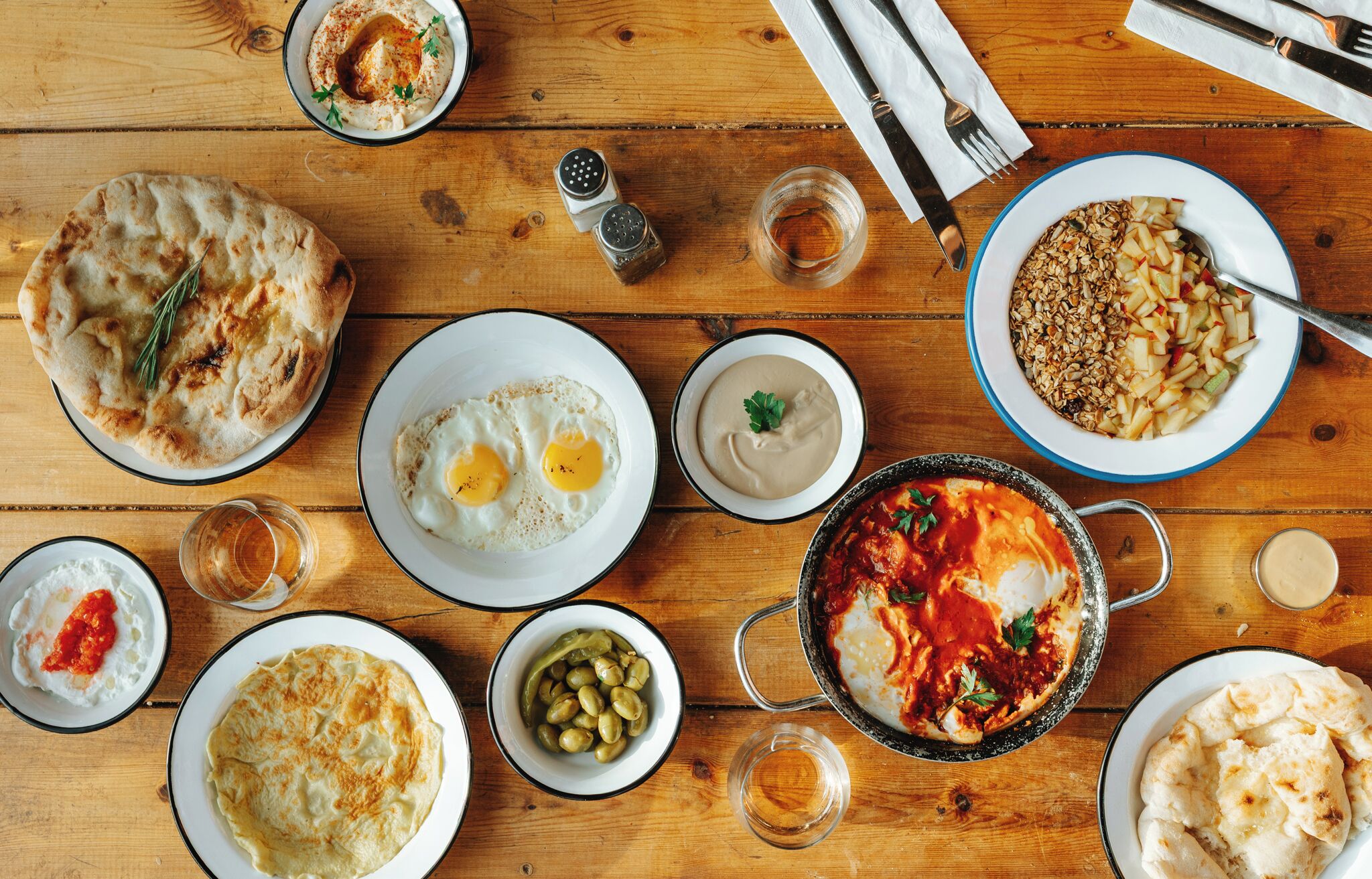 A wooden table with various dishes including flatbread, eggs, olives, and a stew.