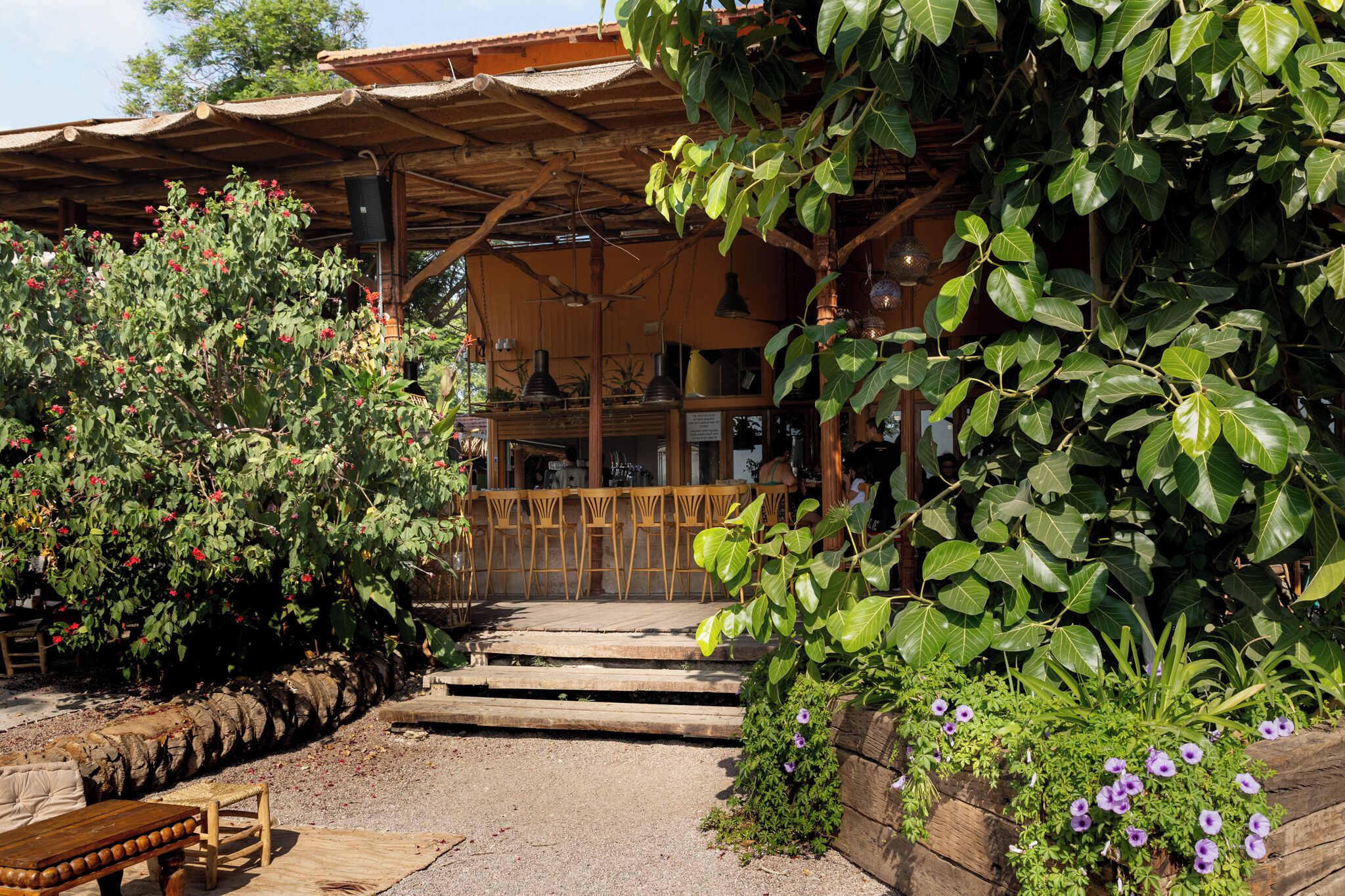 A rustic outdoor seating area with wooden tables and chairs, a stone wall, and a covered wooden structure with a thatched roof.