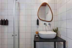 A bathroom with a white tiled wall, a round mirror, a white sink, a black countertop, a potted plant, and toiletries on the wall. - Solaya Kineret (Migdal)