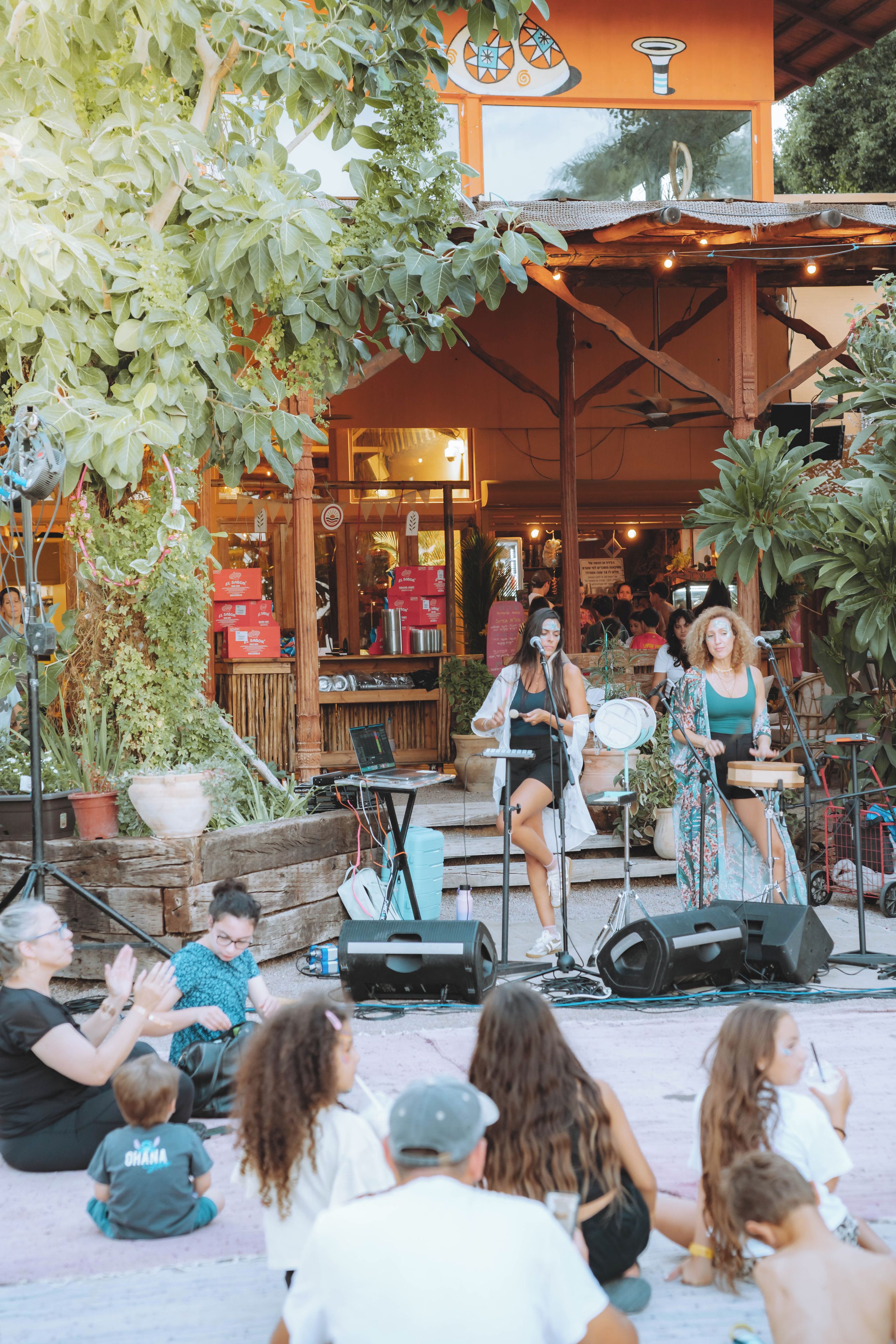 Un escenario al aire libre con músicos actuando, rodeado de un público sentado, y un restaurante al fondo.