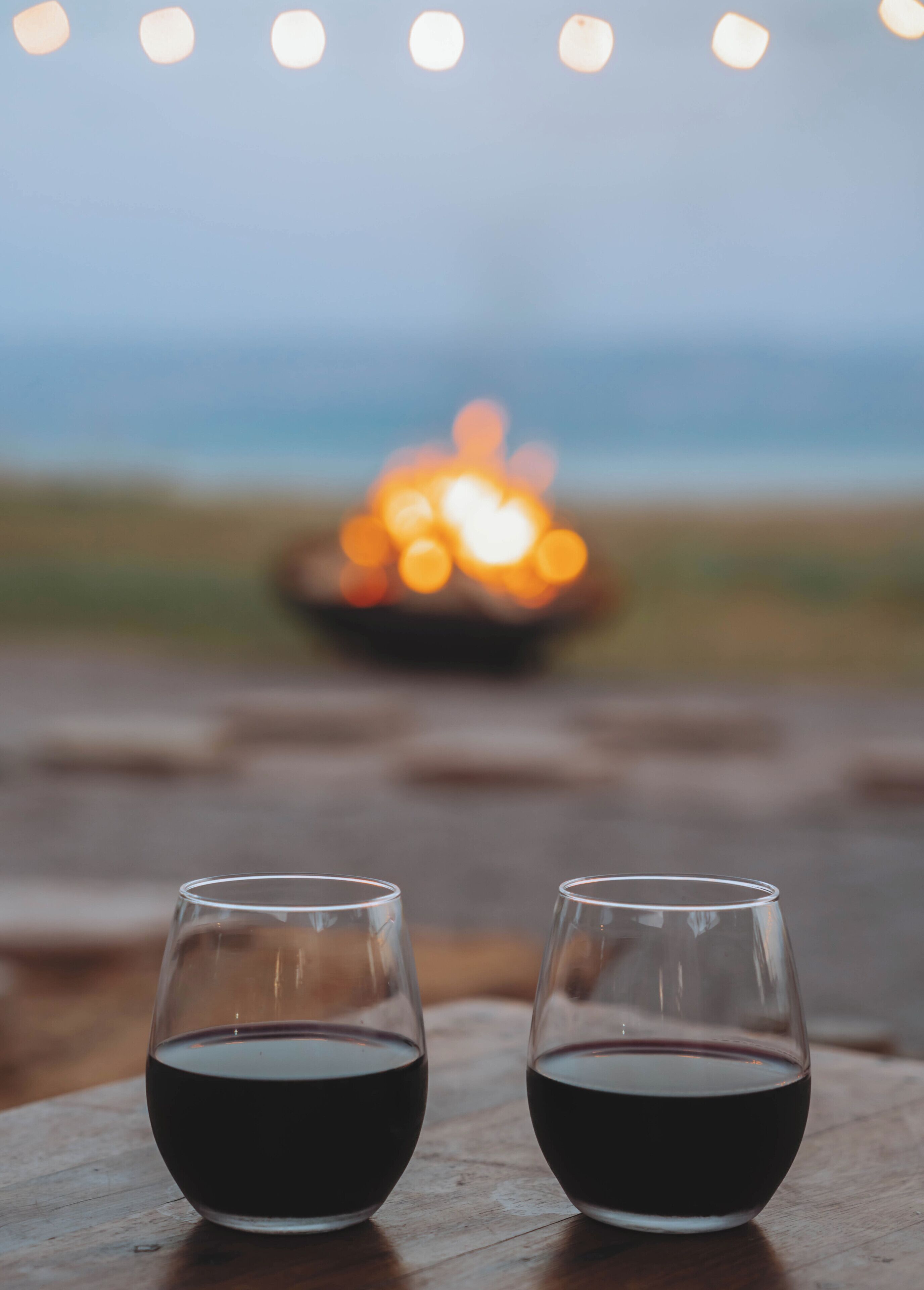 Two glasses of red wine on a wooden table with a blurred background of lights and a beach.