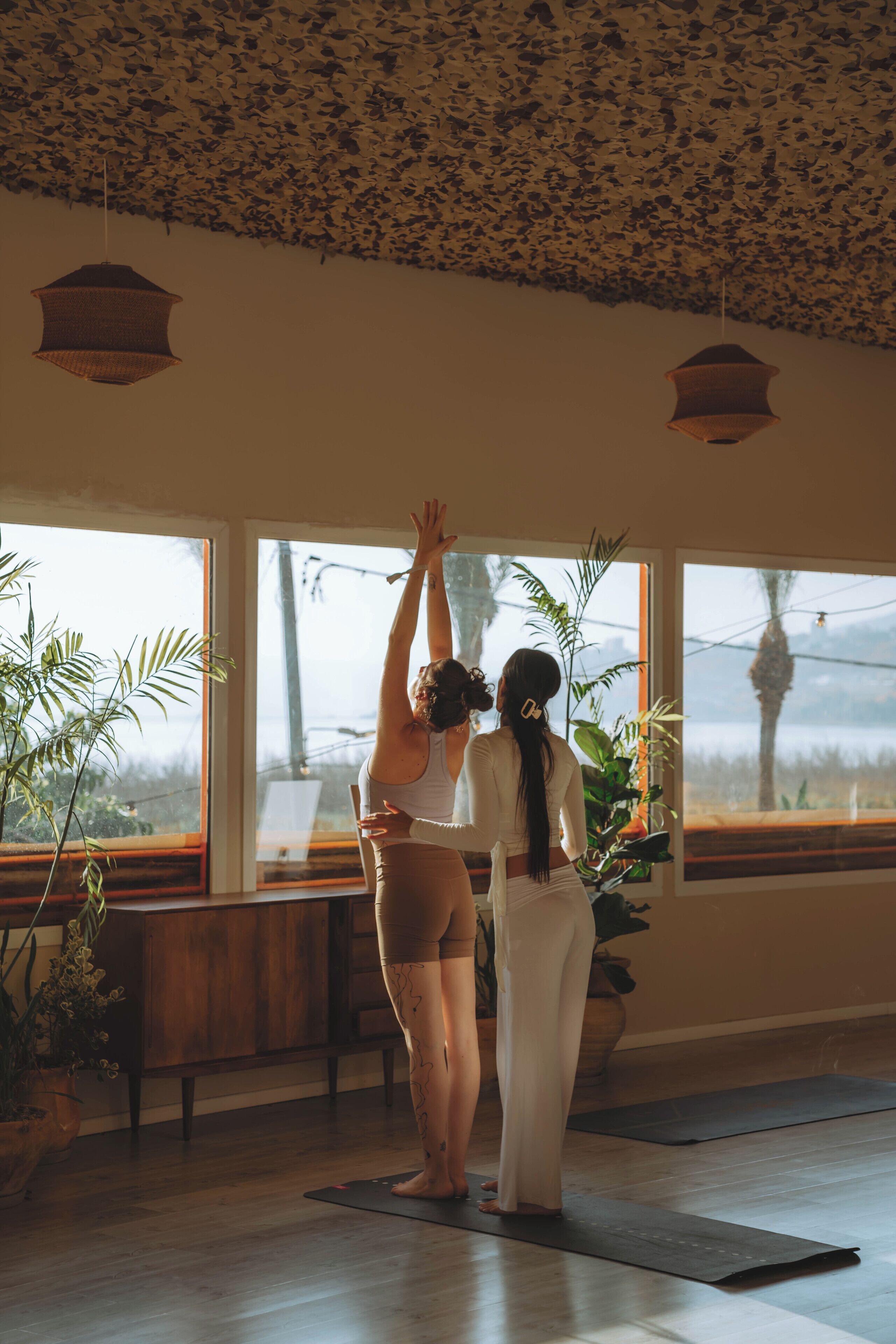 Two people practicing yoga in a room with large windows, wooden floors, and hanging lamps.