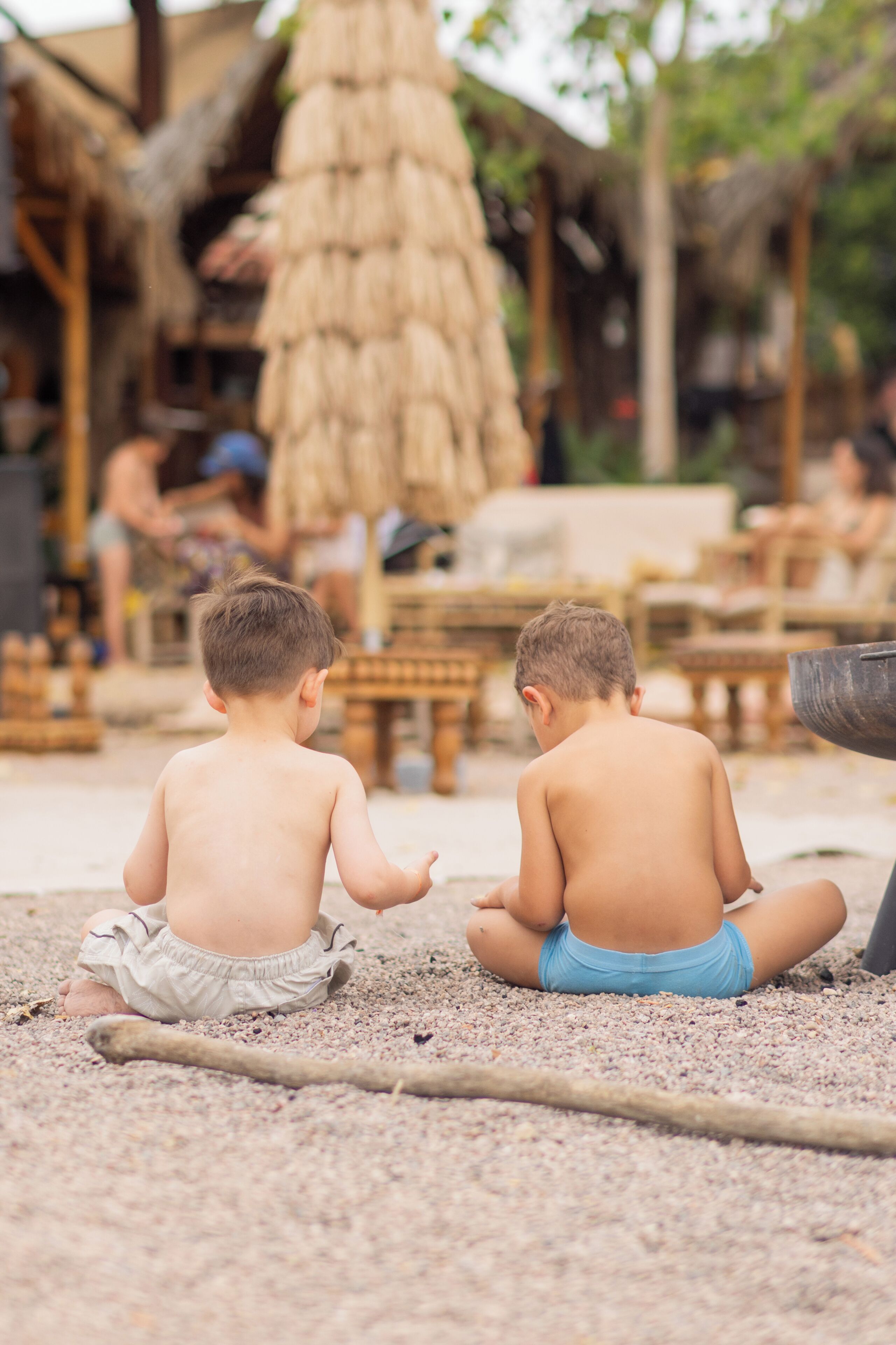 Two children sitting on a sandy surface with a thatched structure and wooden furniture in the background.