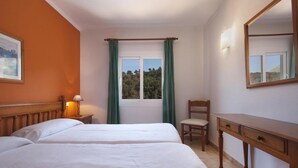 A bedroom with two beds, a wooden dresser, a chair, a nightstand with a lamp, a window with curtains, and a framed picture on the wall. - APT. ALMENDRO Grande, 2 bedrooms with air conditioning, balcony, pool, close to the beach 400 m, WIFI (Cala Sant Vicenç)