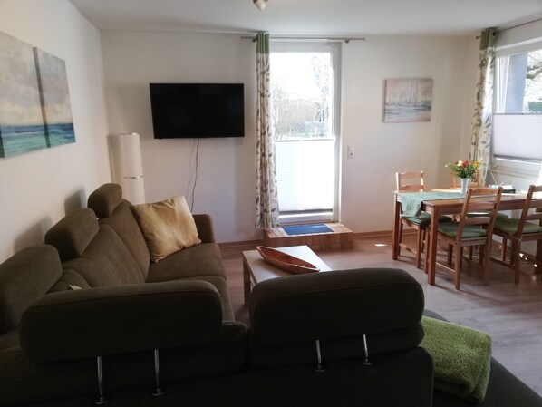 A living room with a sofa, a flat-screen TV mounted on the wall, a dining table with chairs, and a window with curtains.