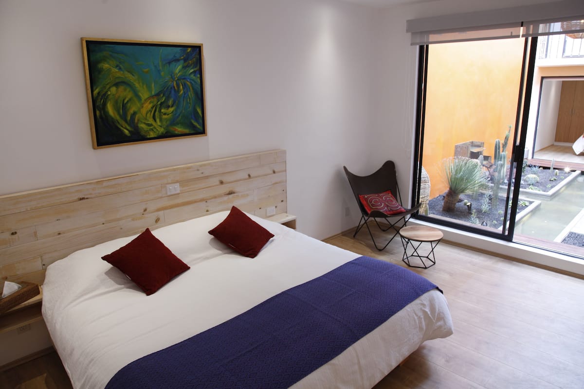 Habitación de hotel con una cama grande, una mesita, una silla y vista a un patio con plantas y piscina.