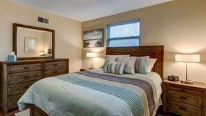 A bedroom with a large bed, a dresser, a nightstand, a window with blinds, a ceiling light, and a painting on the wall.