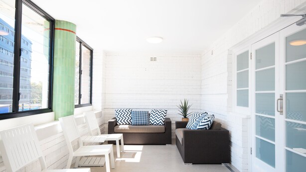 Een zonnig terras met witte rieten meubels, een groene, cilindervormige pilaar en uitzicht op een gebouw door grote ramen.