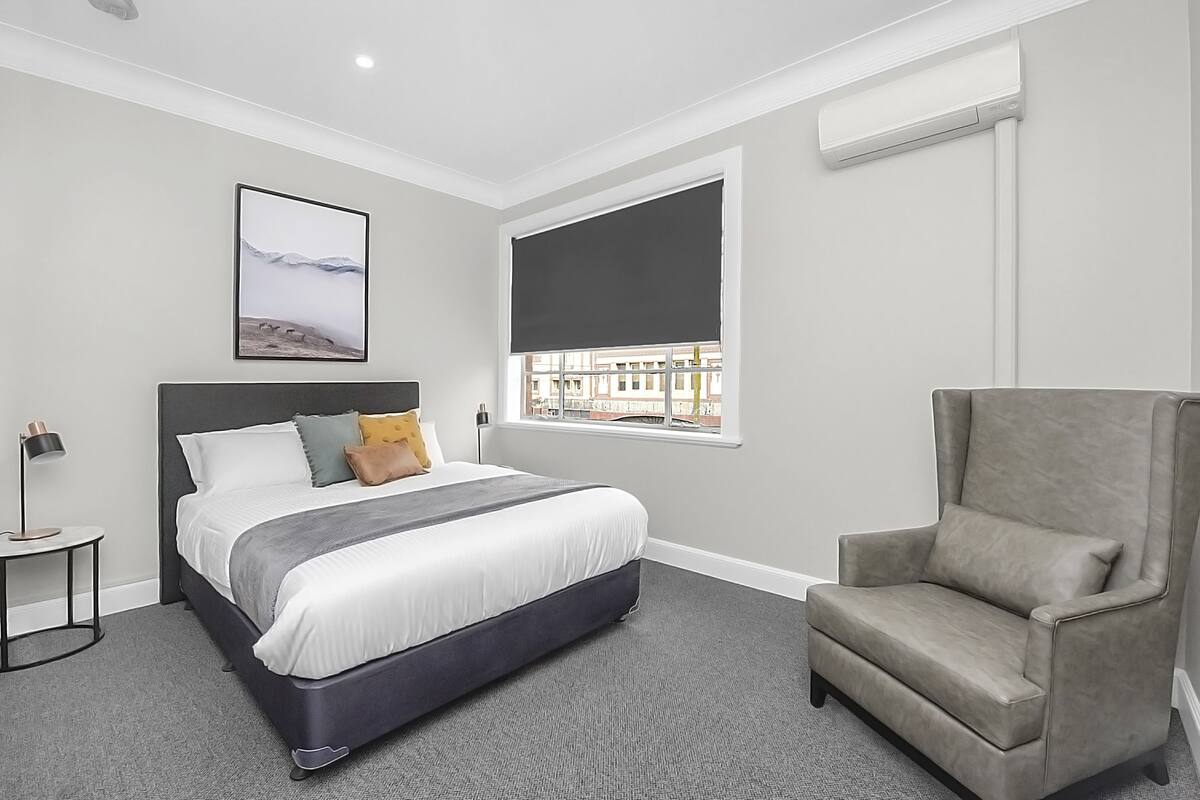 Una habitación de hotel moderna con una cama grande, un sillón gris, un ventanal con vistas a edificios y un aire acondicionado instalado.