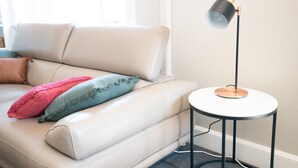 Een witte leren bank met twee kussens, een kleine ronde tafel met een lamp, en een raam op de achtergrond.