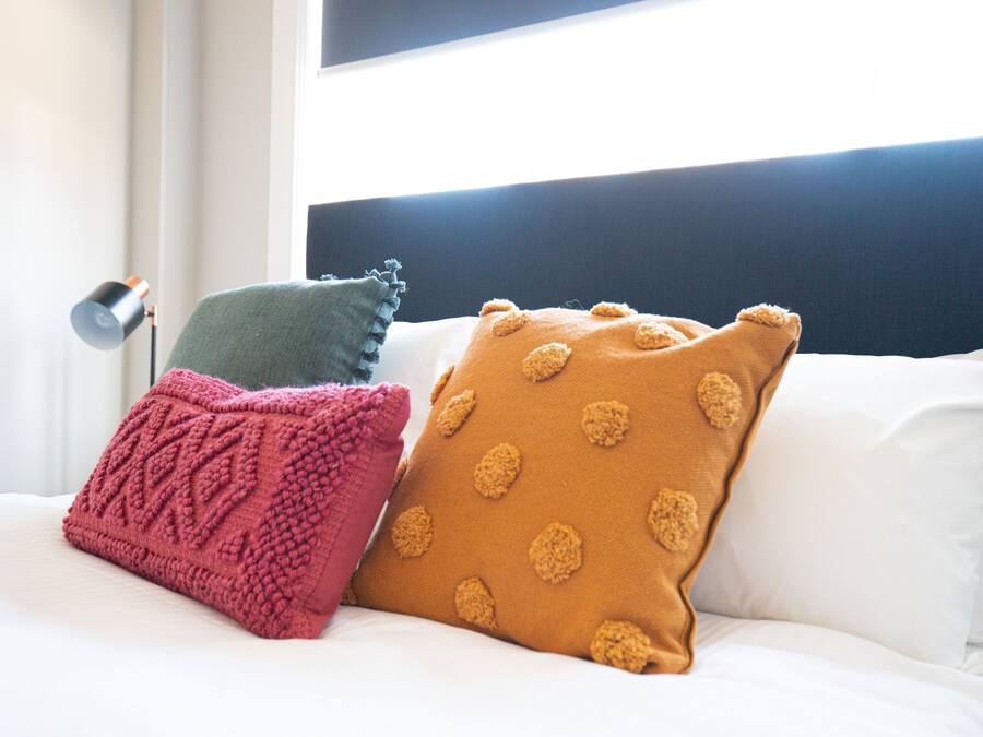 Un letto con biancheria bianca, una testiera nera e tre cuscini di diversi colori e fantasie.