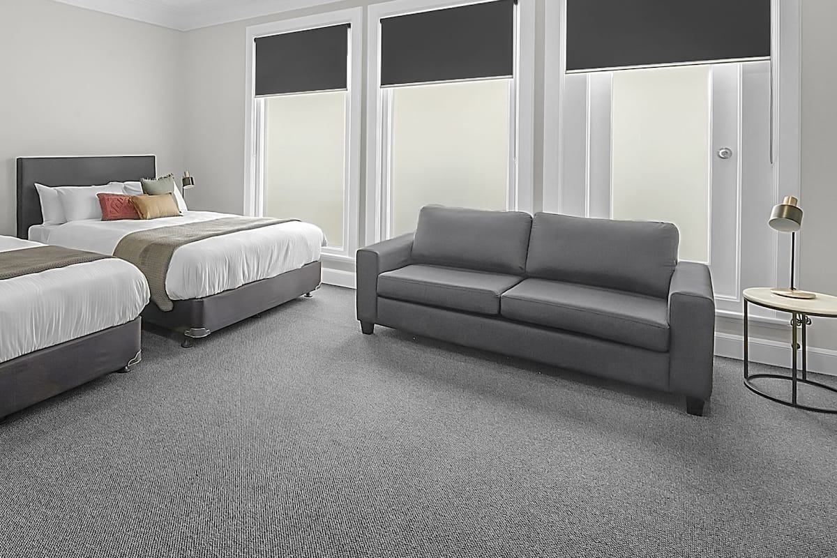 Habitación de hotel con dos camas, un sofá gris, una mesita redonda y ventanas grandes con persianas.