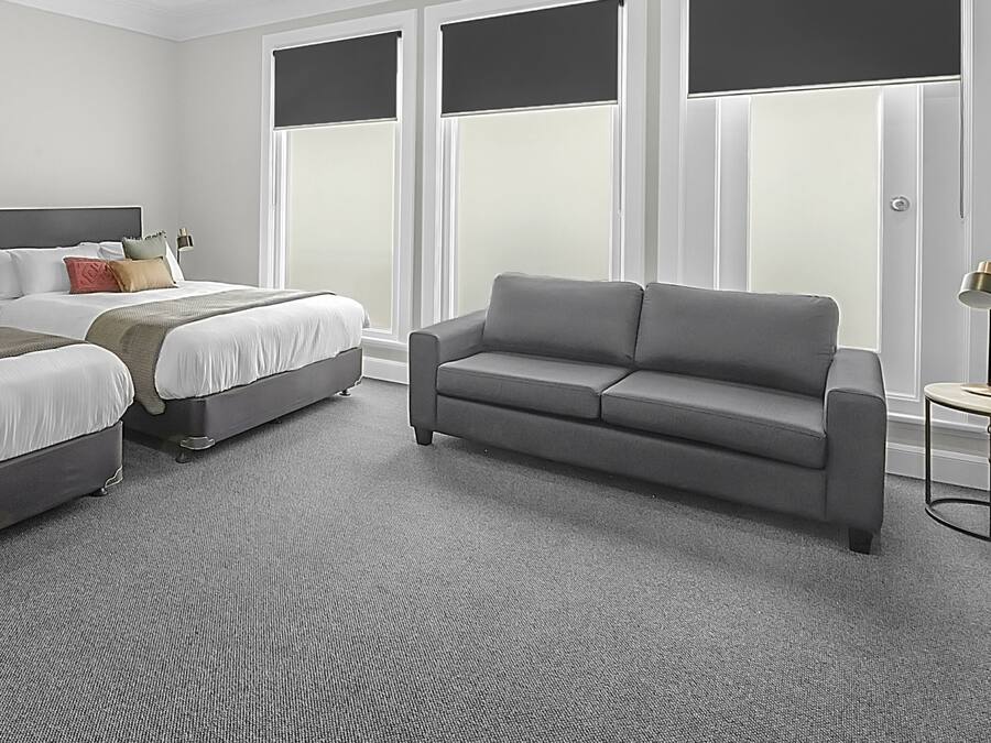 Quarto de hotel com duas camas, um sofá cinzento, uma mesa redonda pequena e grandes janelas com persianas.