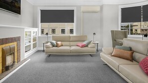 Een moderne woonkamer met een beige bank, een open haard en grote ramen met jaloezieën.