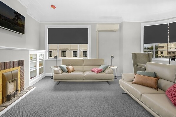 Ein modernes Wohnzimmer mit einem beigefarbenen Sofa, einem Kamin und großen Fenstern mit Jalousien.