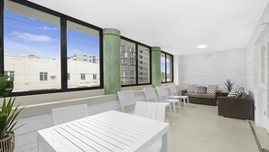 Een moderne zithoek met witte meubels, grote ramen en uitzicht op gebouwen.