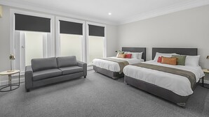 Een hotelkamer met een grijze bank, twee eenpersoonsbedden, een klein tafeltje en een groot raam met jaloezieën.