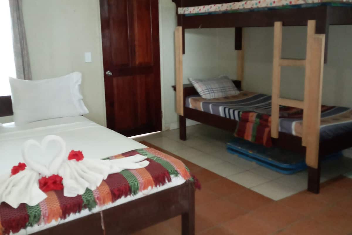 Un dormitorio con una cama, una cama nido de madera, un ventilador de techo, una puerta y piso de baldosas.