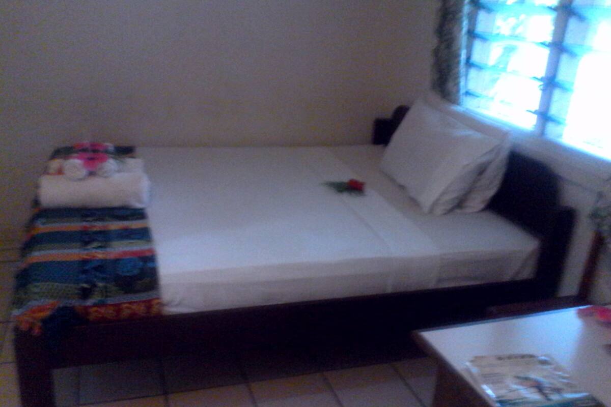 Una cama pequeña y bien hecha con sábana blanca y almohadas, una manta a rayas y una pequeña flor roja. Hay una ventana con persianas entreabiertas y una mesa de madera con una revista encima.