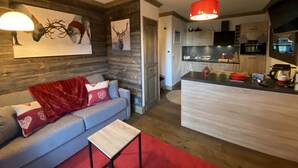 A modern living room with a grey sofa, red throw blanket, and a red rug. There is a wooden wall with deer artwork and a kitchen area with a black countertop and wooden cabinets. - F003 - 40 m2 - 2P - 2/4 pers (Prapoutel)