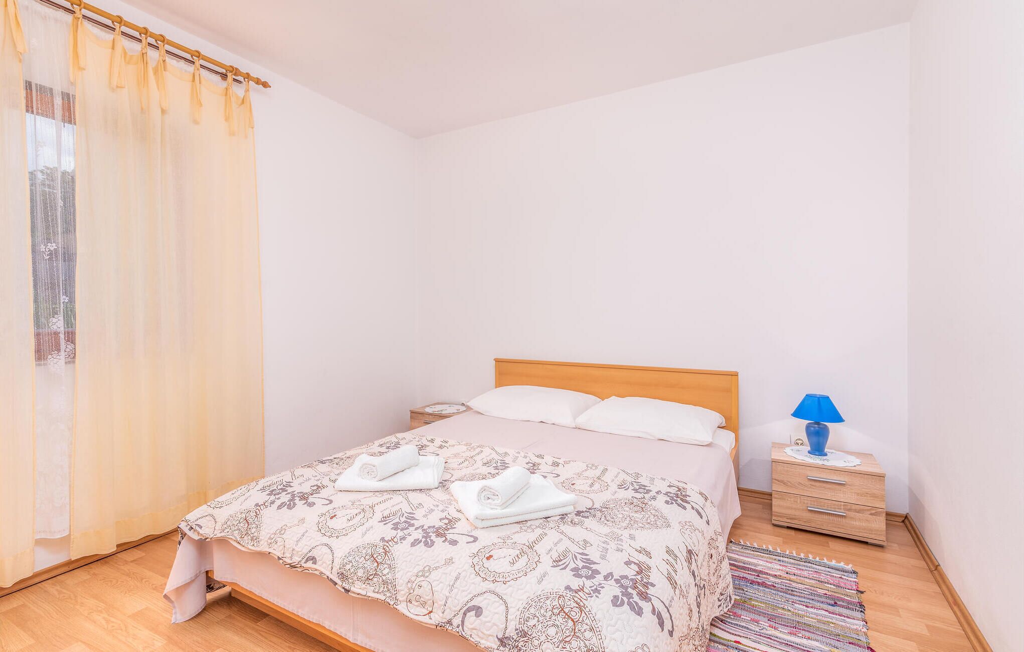 A neatly made bed with a patterned bedspread, a wooden headboard, and two pillows. There is a small bedside table with a lamp and a folded towel.