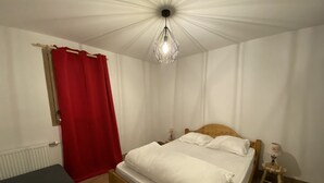 A bedroom with a wooden bed, white bedding, a red curtain, a small bedside table with a lamp, and a wooden bench.