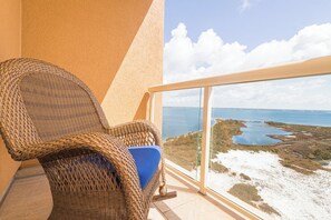Une chaise en osier avec un coussin bleu, placĂ©e sur un balcon donnant sur un paysage cĂŽtier avec de lâeau et de la vĂ©gĂ©tation.