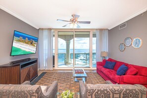 A living room with a television, red sofa, patterned chairs, a ceiling fan, and a view of the ocean through large glass doors.