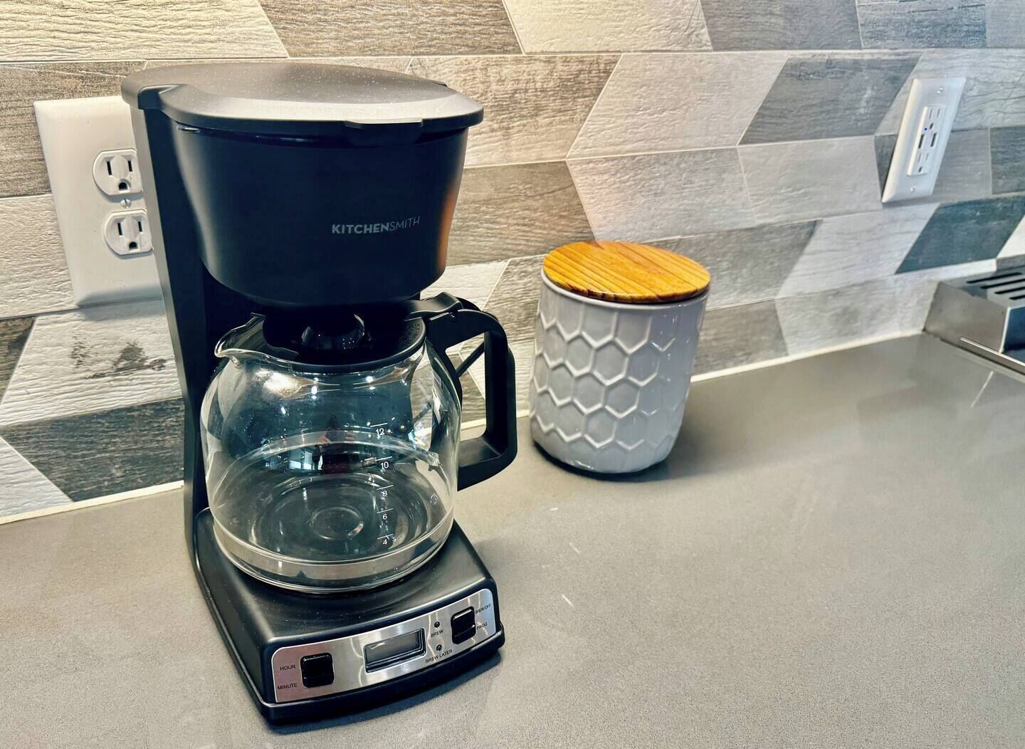 Een koffiezetapparaat op een keukentafel, met een achterwand van hexagonaal patroon tegels en een stopcontact.