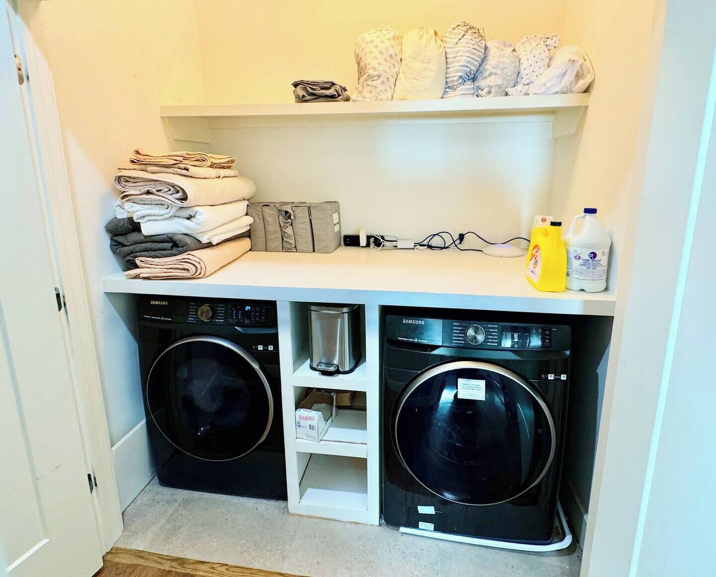 A laundry room with two front-loading washing machines, a shelf with folded towels, a bottle of laundry detergent, and a Samsung device on the counter.