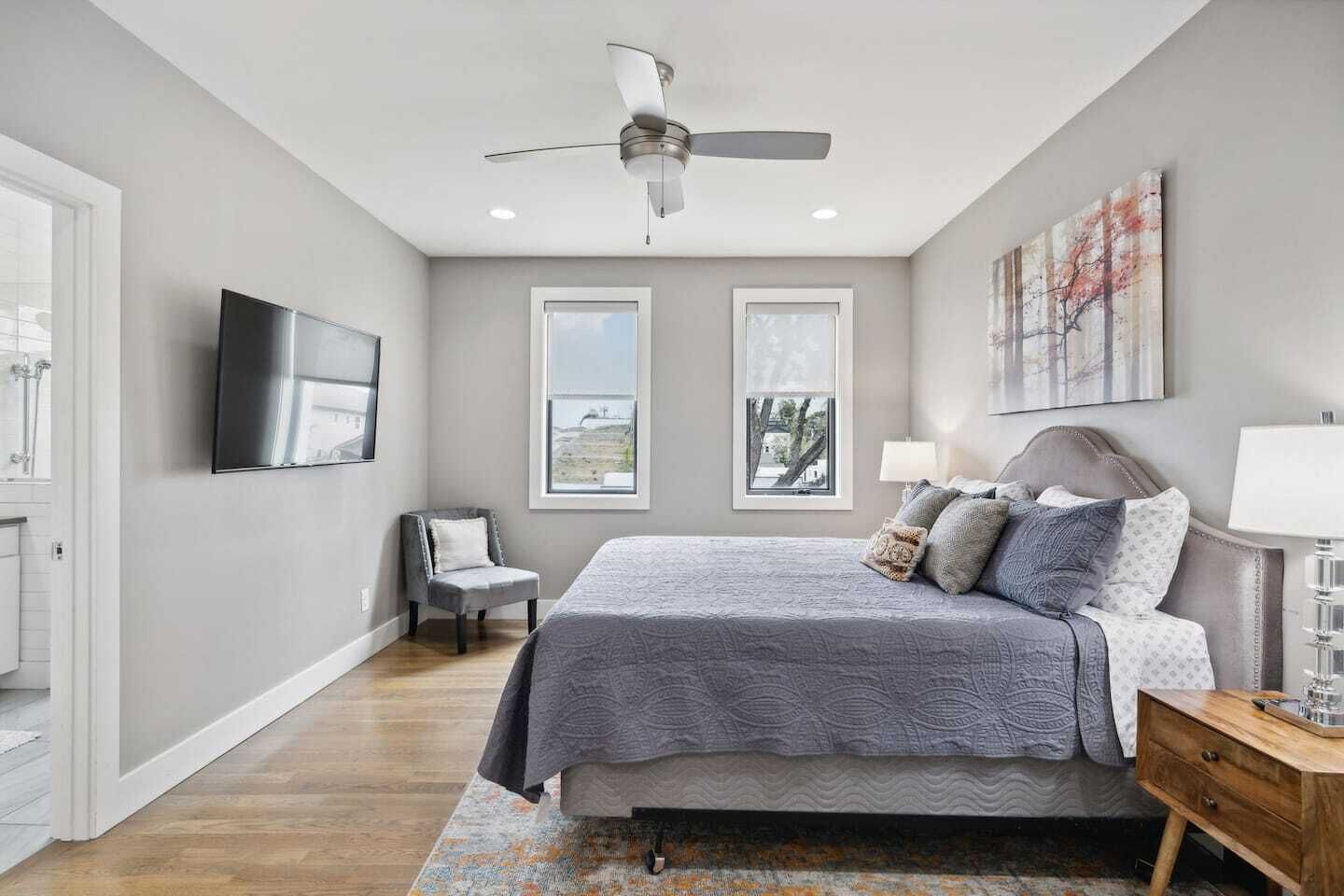 A bedroom with a bed, a chair, a nightstand, a lamp, a ceiling fan, and two windows.