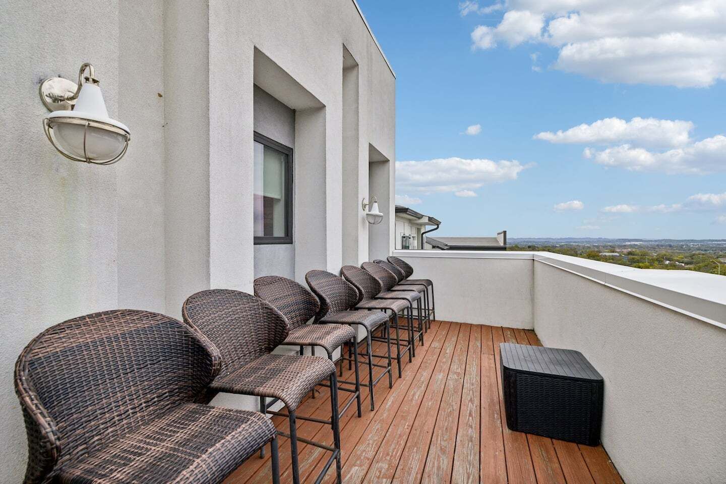 A balcony with wicker chairs and a black ottoman, a white wall with a window, and a clear view of the landscape.
