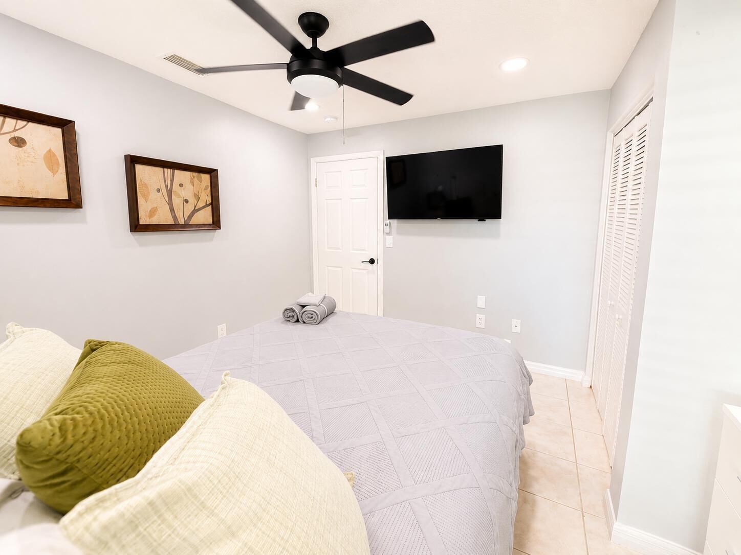 A bedroom with a bed, pillows, a ceiling fan, a door, and a TV on the wall.