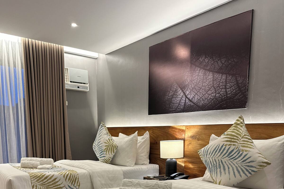 A hotel room with two beds, a large window with curtains, a wall-mounted air conditioner, and a large framed artwork above the beds.