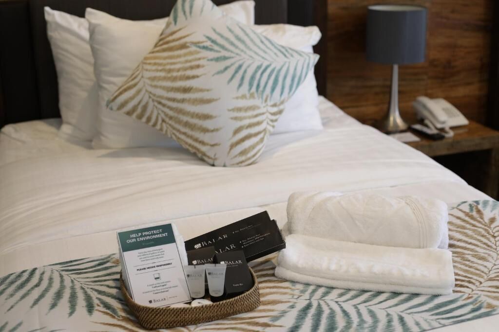 Un lit bien fait, agrémenté d’un coussin orné de motifs de feuilles, d’une serviette blanche et d’un panier contenant des articles de toilette et une carte encourageant la protection de l’environnement.
