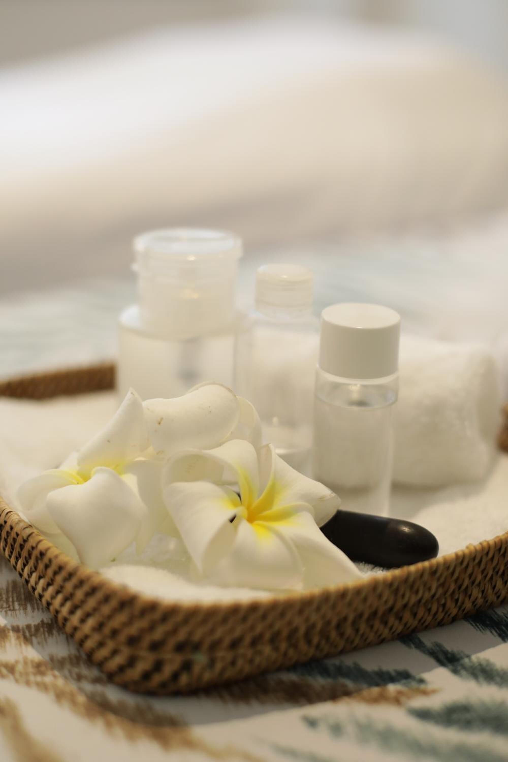 A tray with a woven basket containing white flowers, a black spoon, and several small bottles, likely containing toiletries, on a patterned fabric surface.