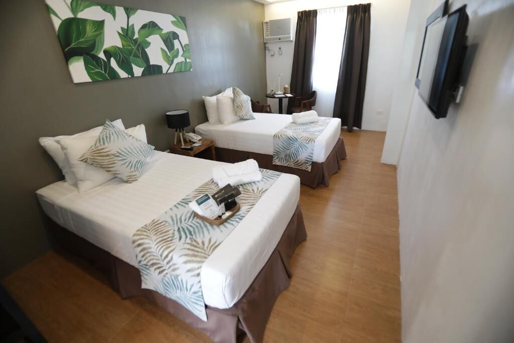 A hotel room with two beds, a small table with a lamp, a TV mounted on the wall, and a large window with curtains.