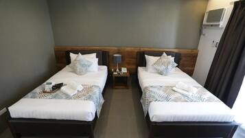 A hotel room with two single beds, each with a wooden headboard, white bedding, and decorative pillows. There is a nightstand with a lamp between the beds, and a window with curtains on the right side.