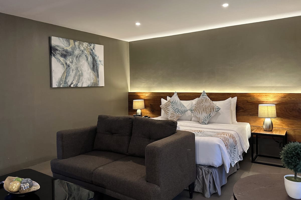 A modern hotel room with a large bed, a sofa, a small table, and a potted plant.