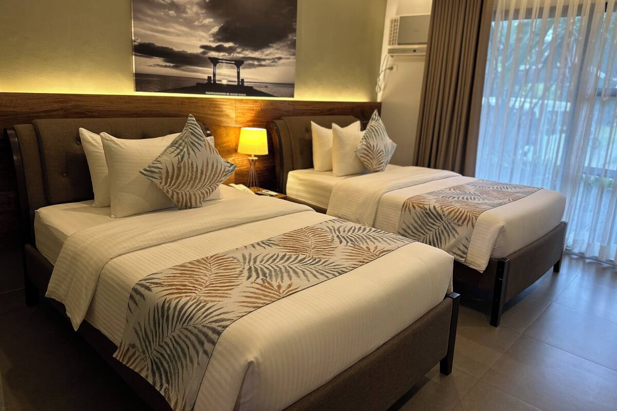 A hotel room with two beds, a large painting on the wall, a lamp, and a view of the outdoors through the window.