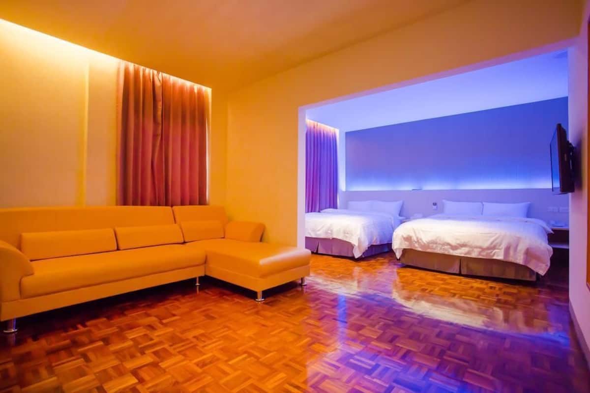 A modern hotel room with a wooden parquet floor, a yellow sofa, two beds with white linens, and a wall-mounted TV.