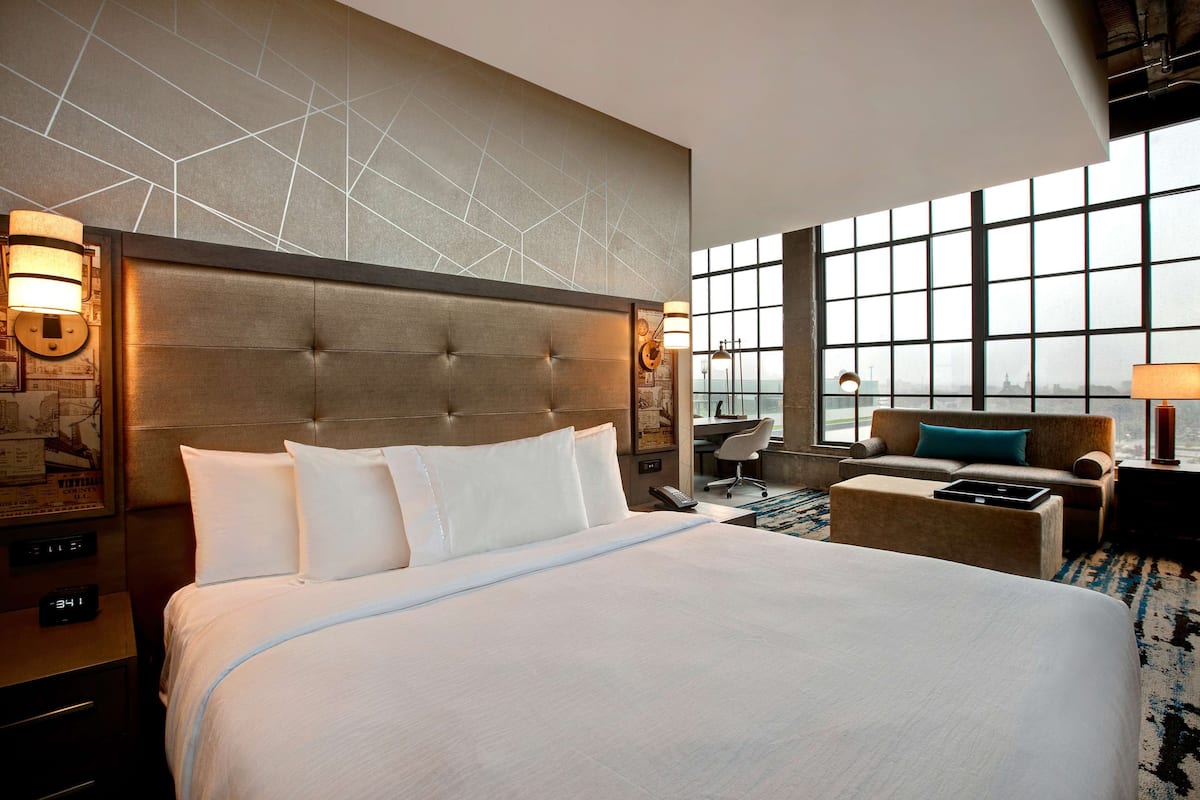 Habitación de hotel con una cama grande, cabecera con diseños geométricos, sofá, escritorio y una ventana amplia con vistas a la ciudad.