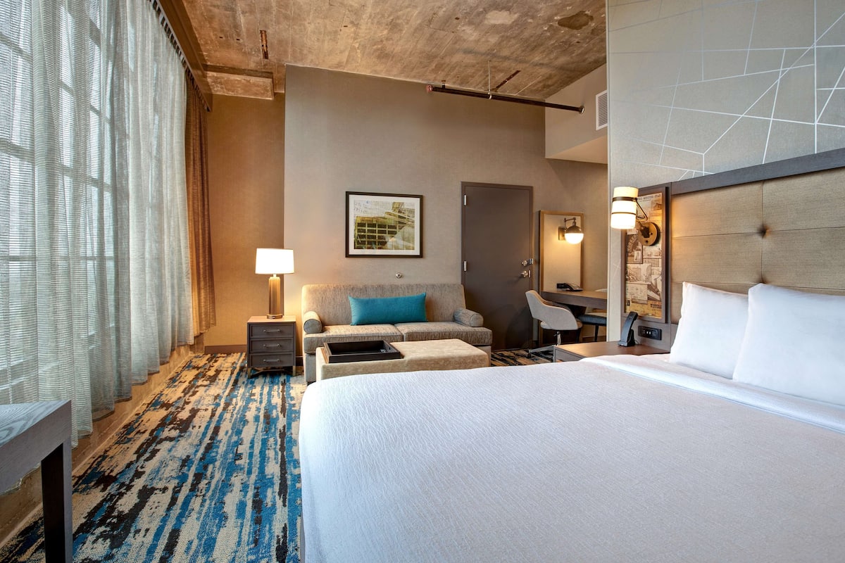 Habitación de hotel con una cama grande, un sofá, un escritorio y una silla. Cuenta con una ventana con cortinas, una lámpara y un cuadro en la pared.